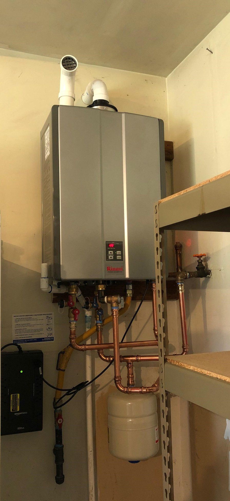 A wall-mounted, silver tankless water heater with copper pipes, a white exhaust pipe, and an expansion tank