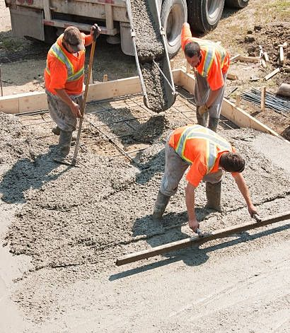 Construction workers in orange vests spreading fresh concrete on a road.
