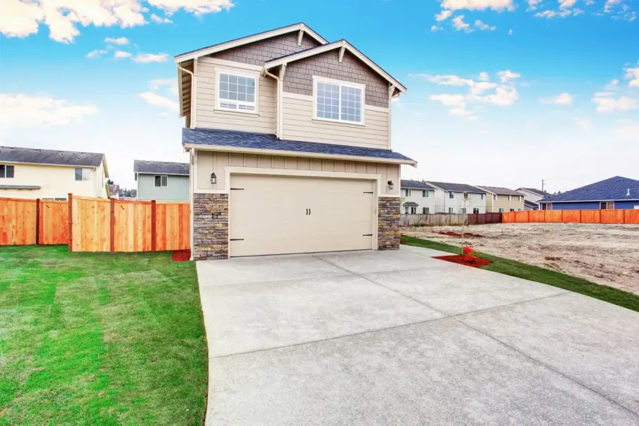 Two-story beige house with a garage, wide driveway, and fenced yard on a sunny day