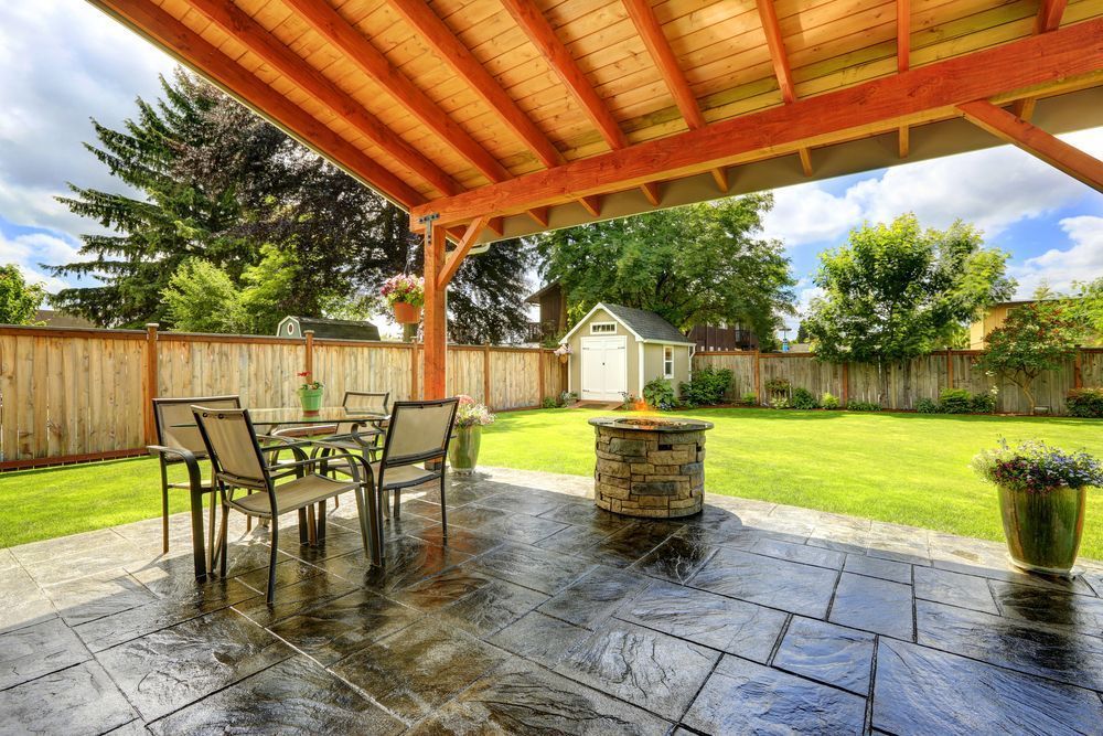 Covered backyard patio with stone floor, metal table and chairs, fenced lawn, and a small garden shed.