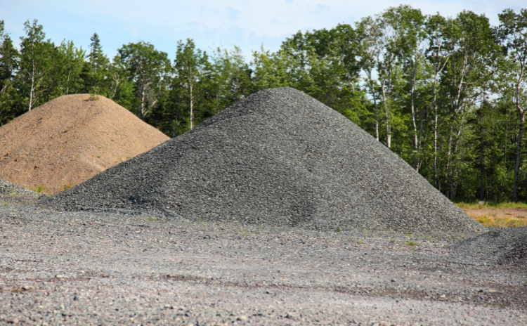 Two piles of aggregate material—one tan and one dark gray—situated in an outdoor area near a line of green trees.