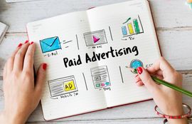 Notebook with “Paid Advertising” sketches, charts, and hands holding a pencil and phone on a desk