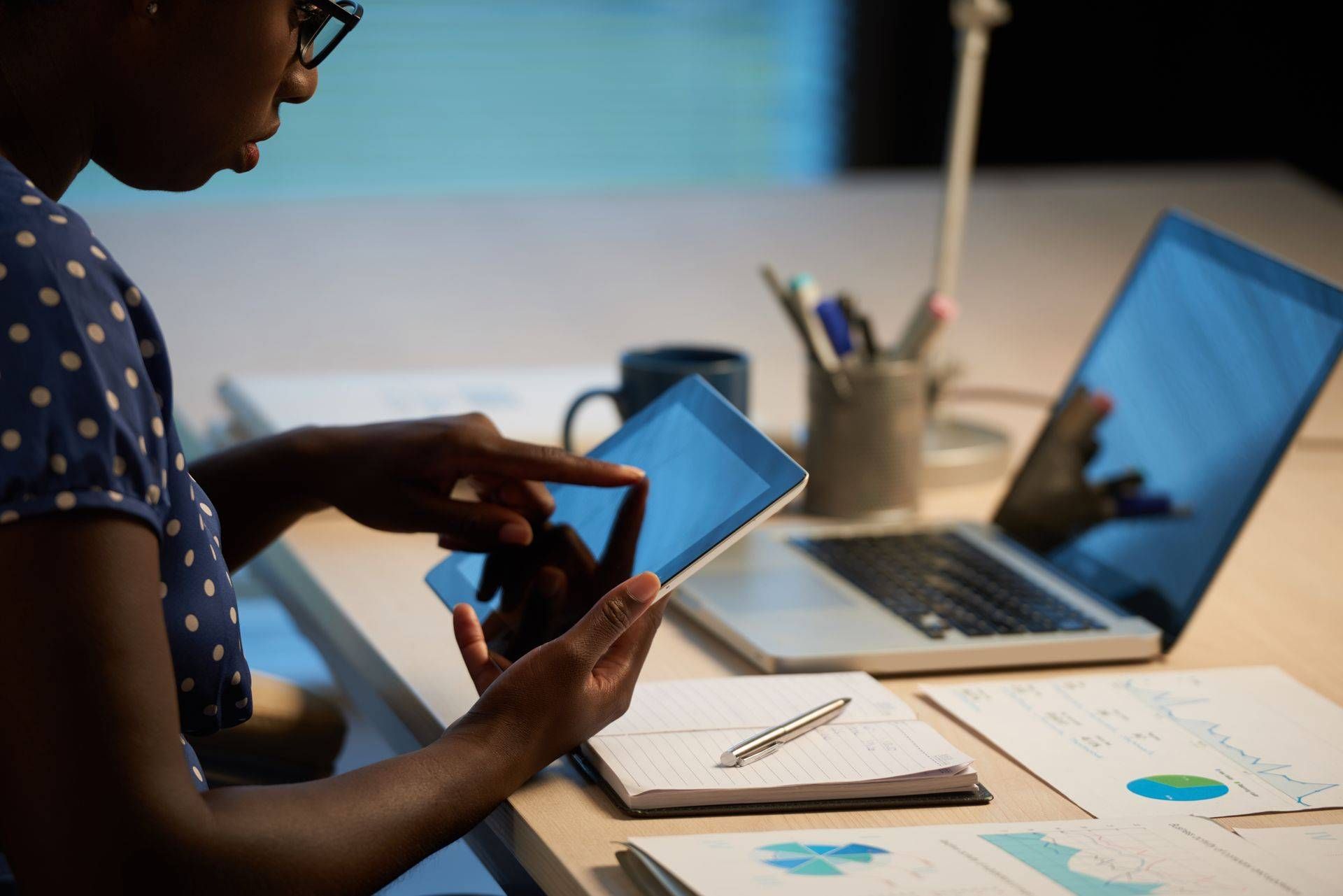 Person using a smartphone beside a laptop at a desk with papers and a notepad