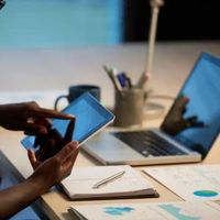 Person using a smartphone beside a laptop at a desk with papers and a notepad