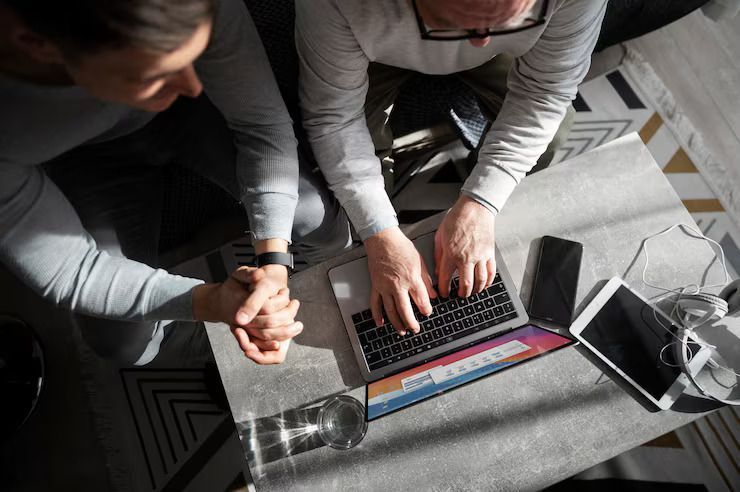 Three people collaborating around a laptop on a gray table, with papers and a glass nearby.