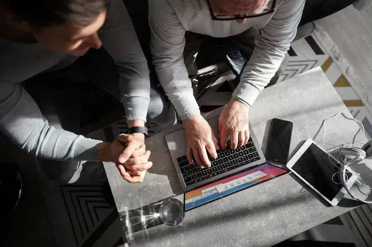 Two people leaning over a laptop on a table, working with papers and a smartphone nearby.