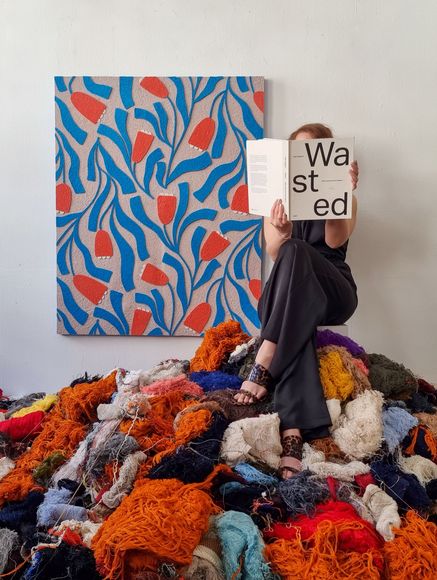 Artist sitting on a pile of colourful textile waste, with a floral wall art panel made from recycled textiles 