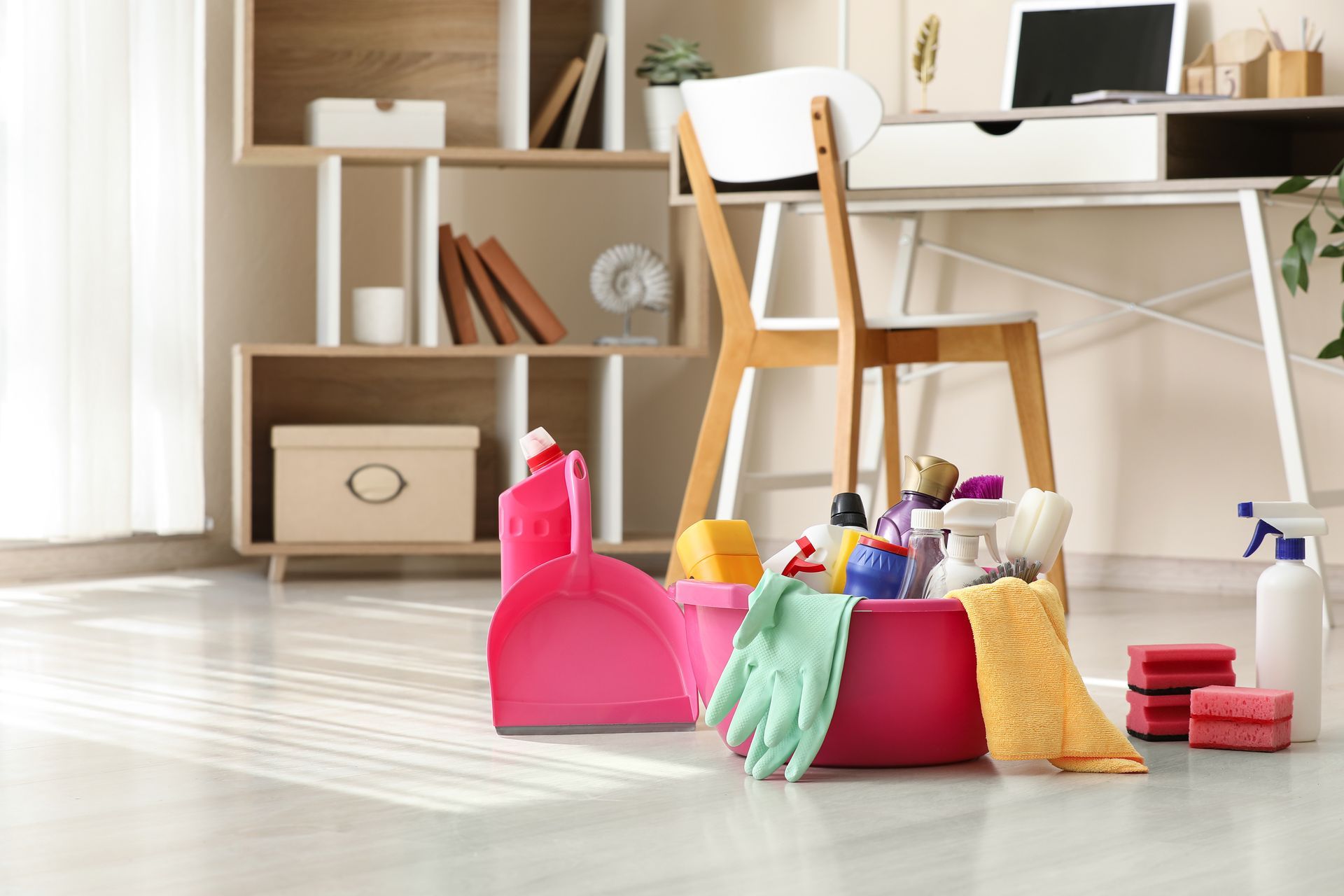 Cleaning supplies in a bright room, with a pink dustpan, gloves, spray bottles, and cloths on the floor Cleaning supplies in a bright room, with a pink dustpan, gloves, spray bottles, and cloths on the floor