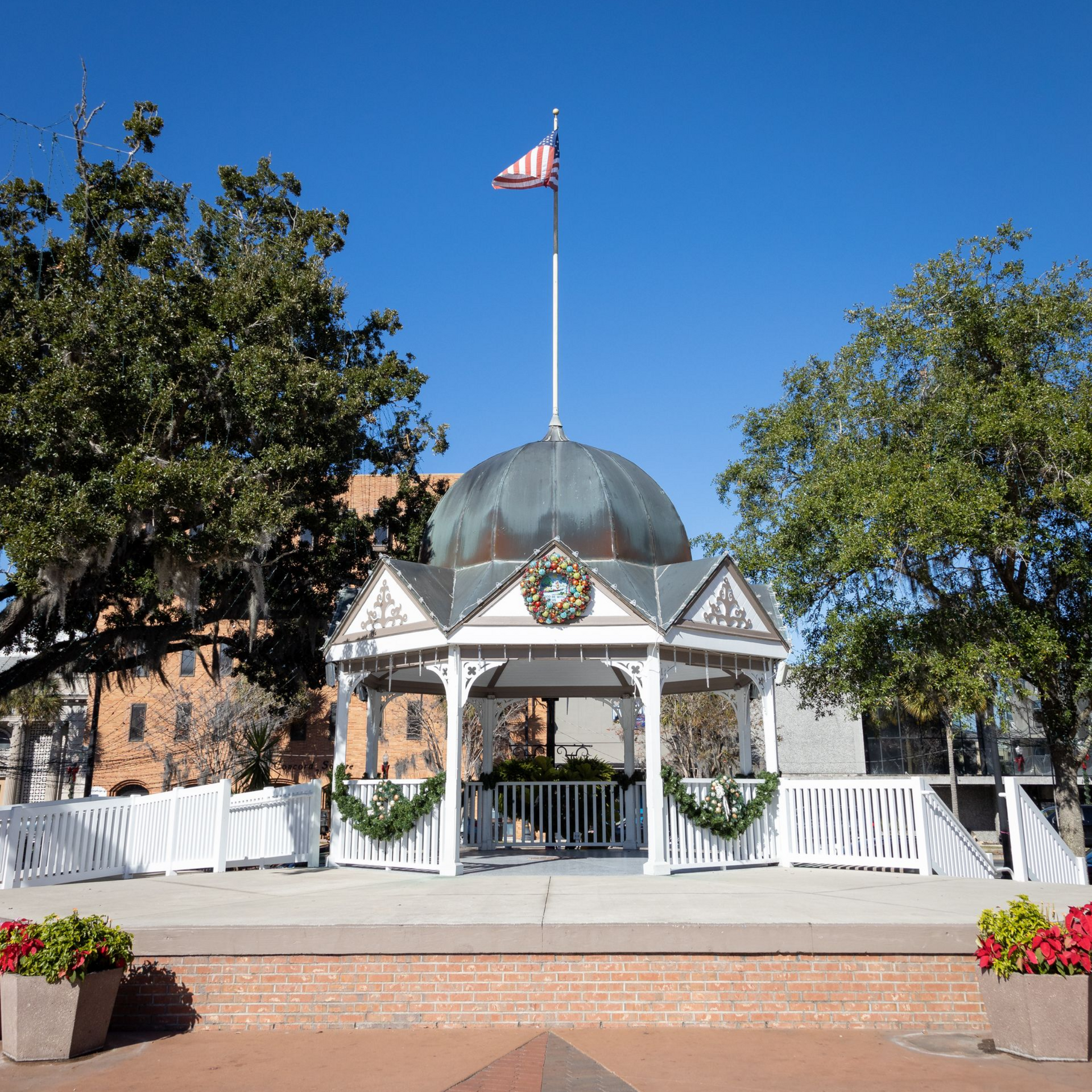 White gazebo with U.S. flag and wreath in a sunny park, framed by trees and flowers.