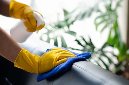 Yellow-gloved hands wipe a black couch with a blue cloth and spray cleaner
