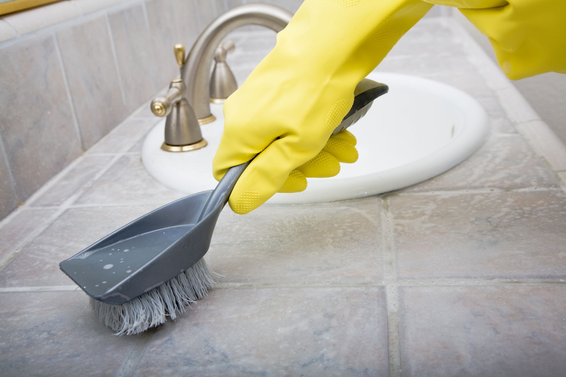 Gloved hand scrubbing a bathroom sink with a brush and dustpan on tiled countertop