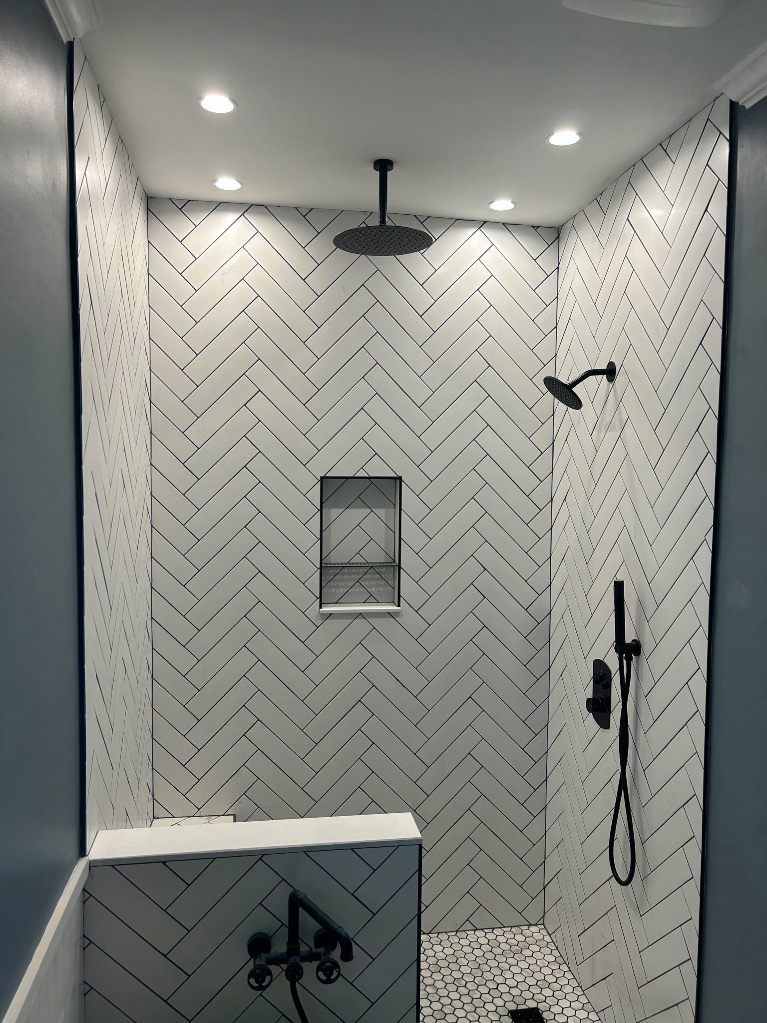 A shower with white herringbone-patterned tile, a dark rainfall showerhead, handheld sprayer, and recessed wall niche.