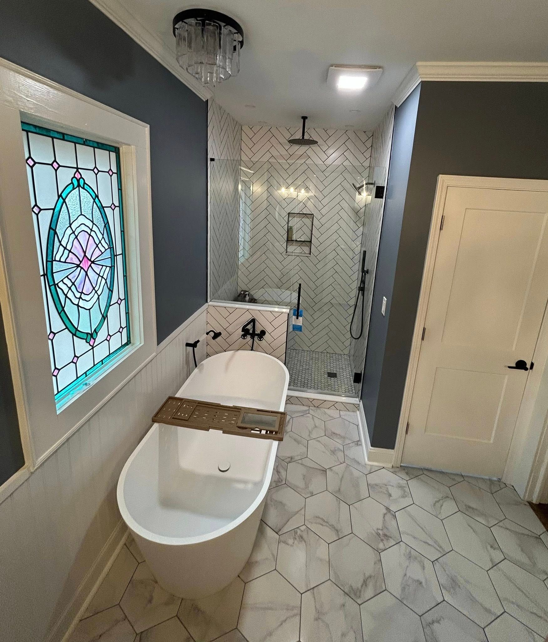 A bathroom featuring a freestanding white bathtub, a stained-glass window, a glass shower, and patterned marble flooring.