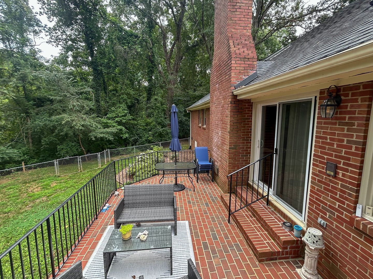 A brick patio with outdoor furniture sits beside a brick house featuring a chimney and glass sliding doors overlooking woods.
