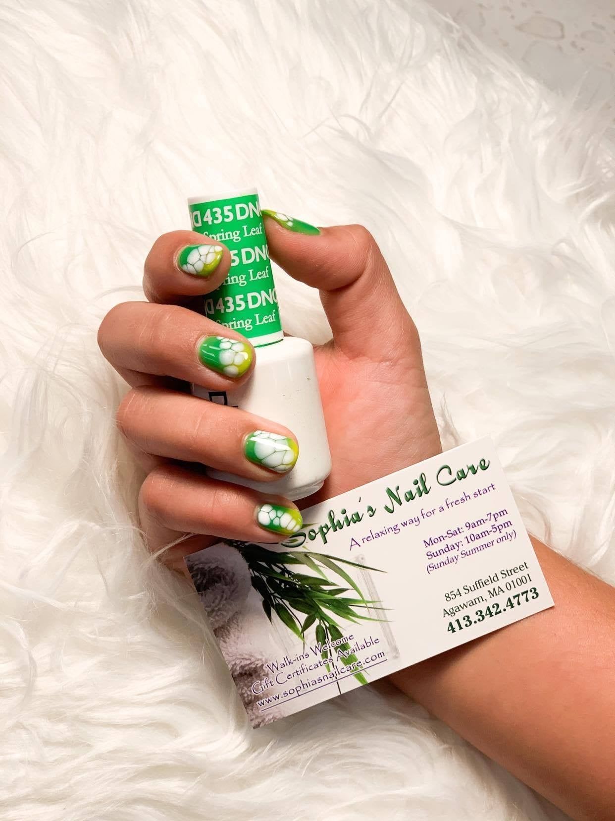 a woman 's hand is holding a bottle of nail polish and a business card for sophie 's nail care . Her nails are painted with green and white nail art.