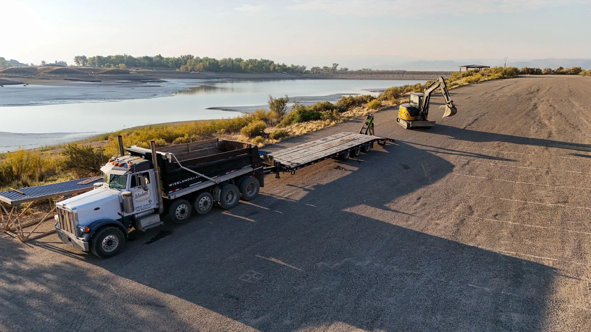 Dump truck and excavator building a gravel road beside a calm river at sunset