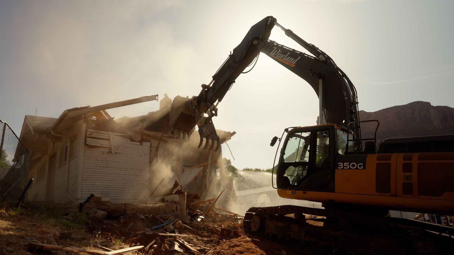Excavator demolishing a building, dust flying in a sunlit construction site.