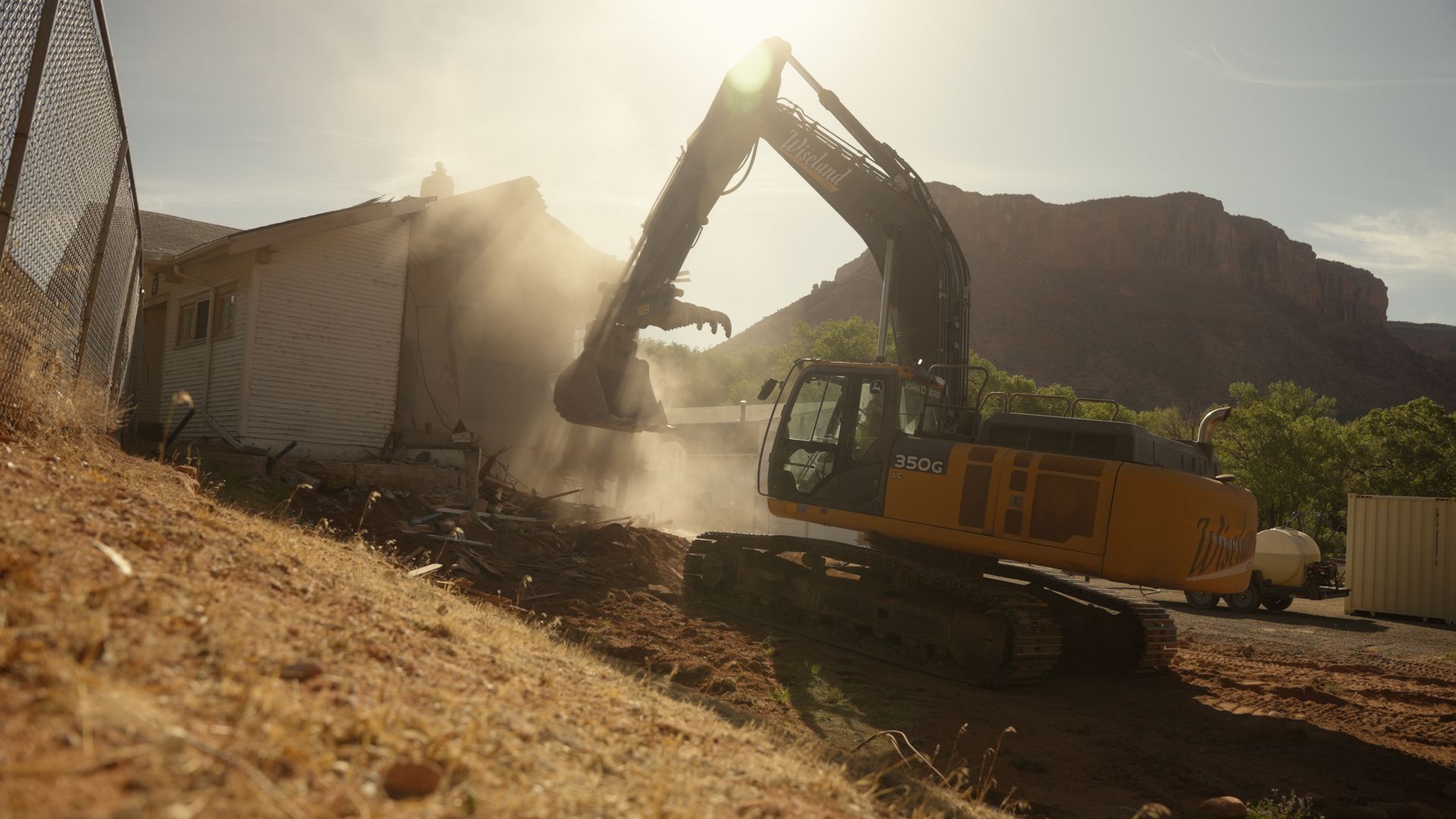 Yellow excavator digging on a dusty hillside beside a building, with sunlight and dust in the air