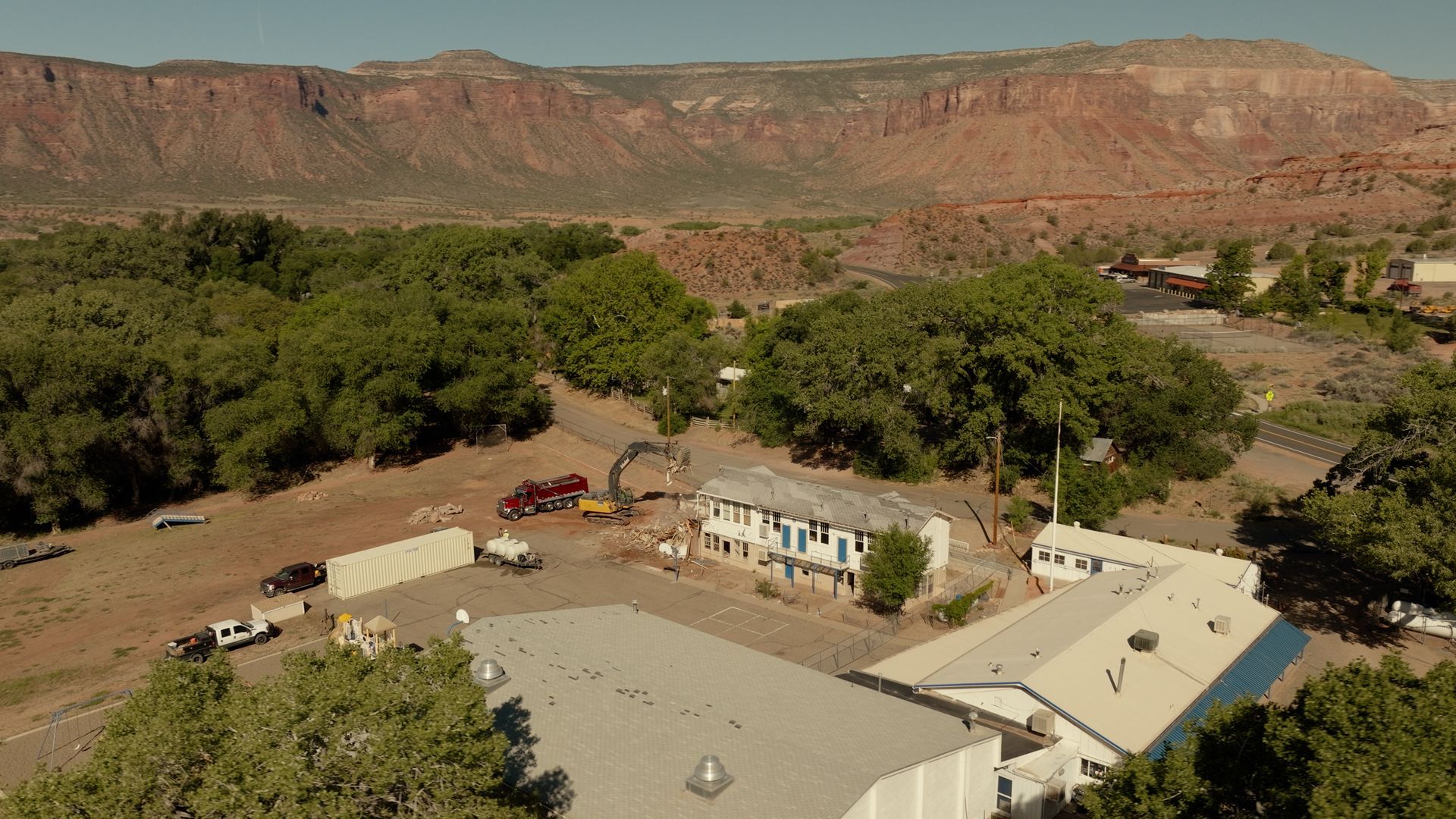 Rural homestead with flat-roof buildings and trees in a dry valley, backed by red cliffs and open land