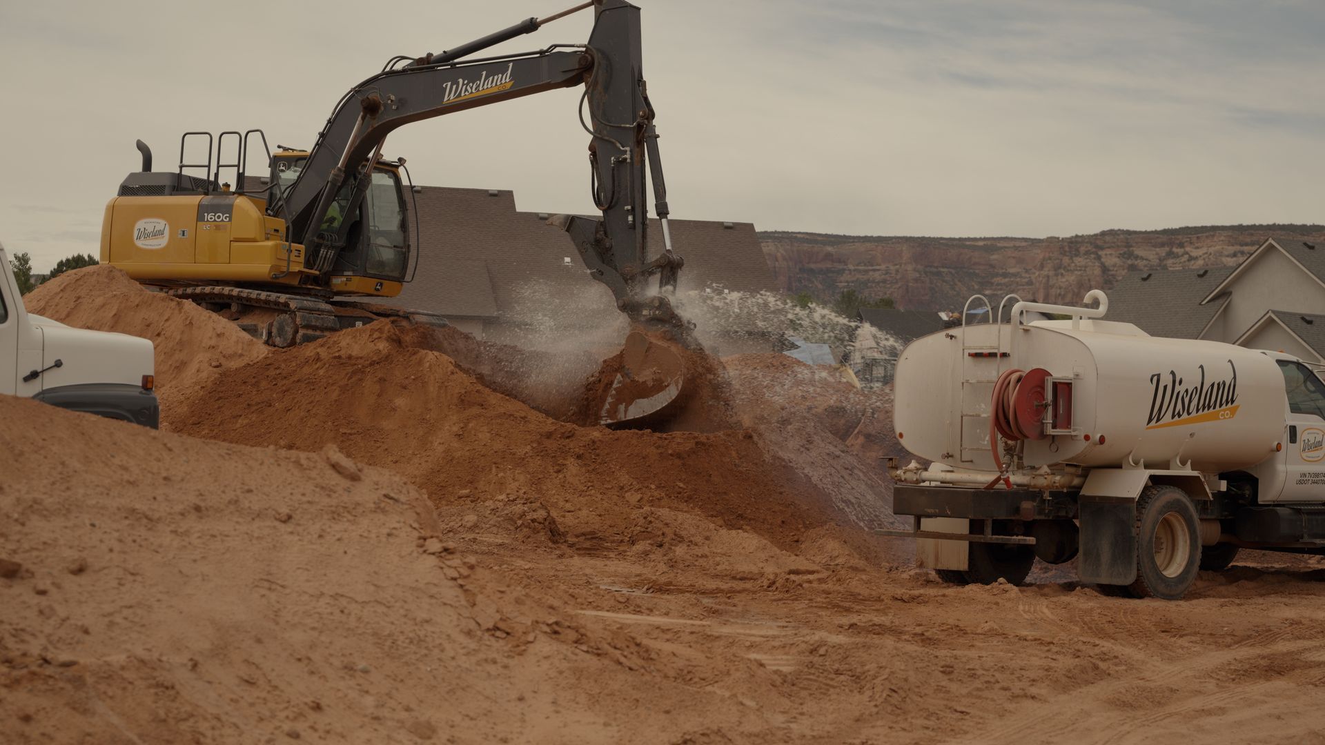 Yellow excavator loading sand into a tanker truck at a dusty construction site.