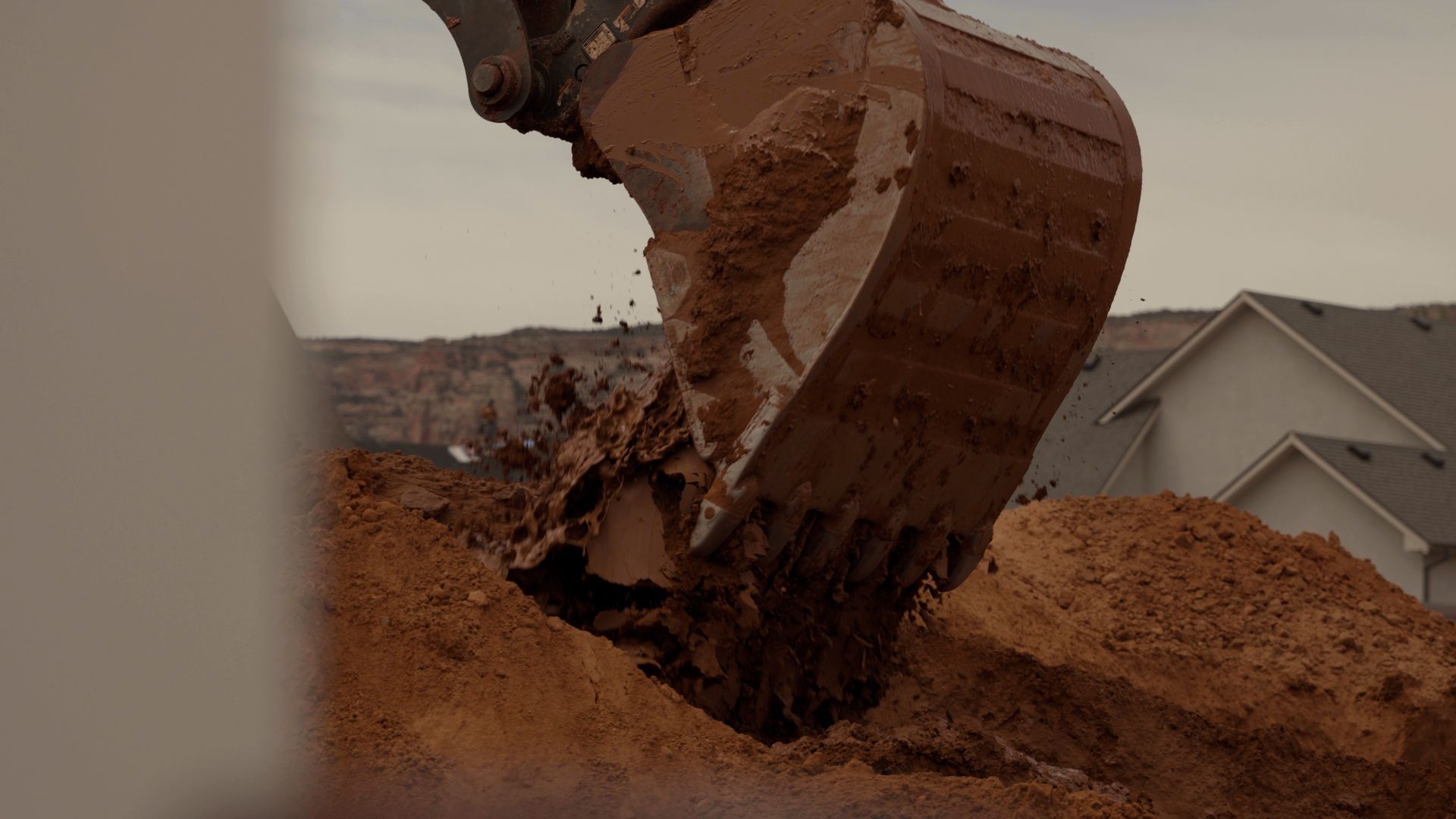 Excavator bucket dumping red dirt at a construction site beside a house