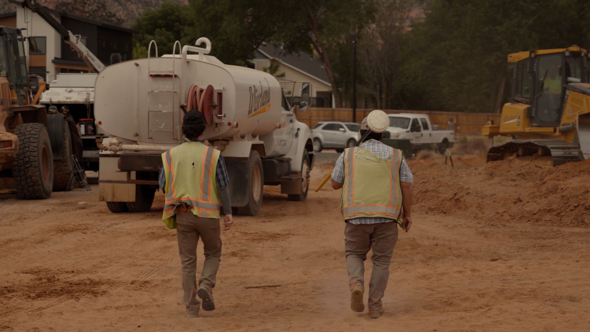 Two workers in safety vests walk toward construction vehicles on a muddy worksite.