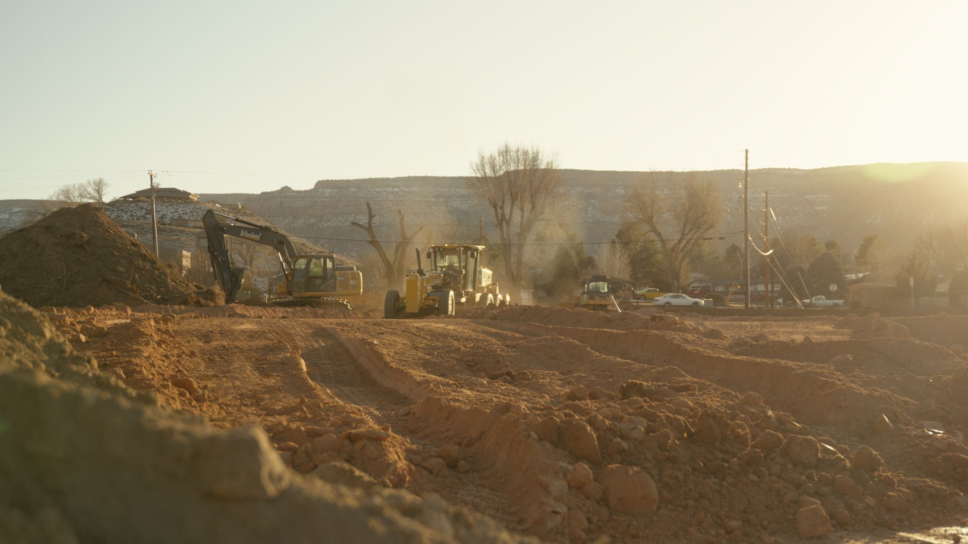 Bulldozers working on a dusty construction site at sunset, with hills in the background