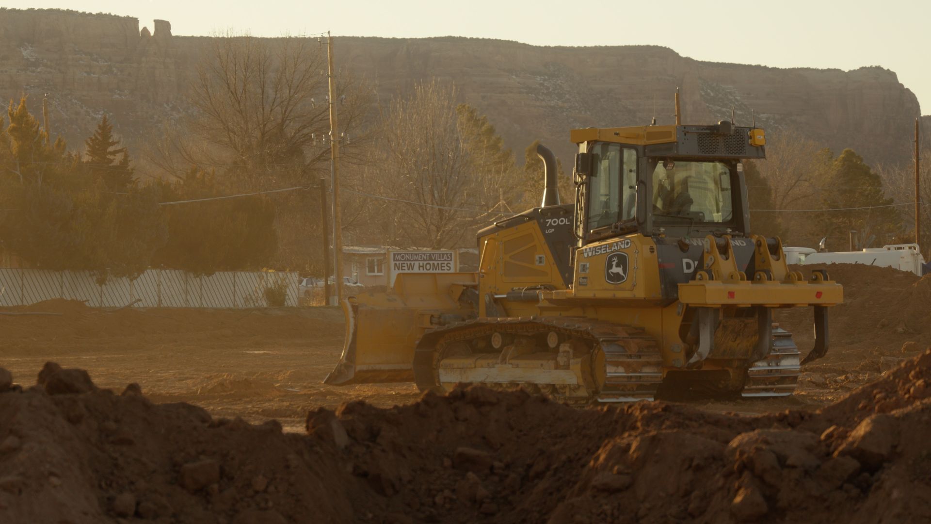 Yellow bulldozer grading dirt on a construction site at sunset, with a dusty hillside in the background
