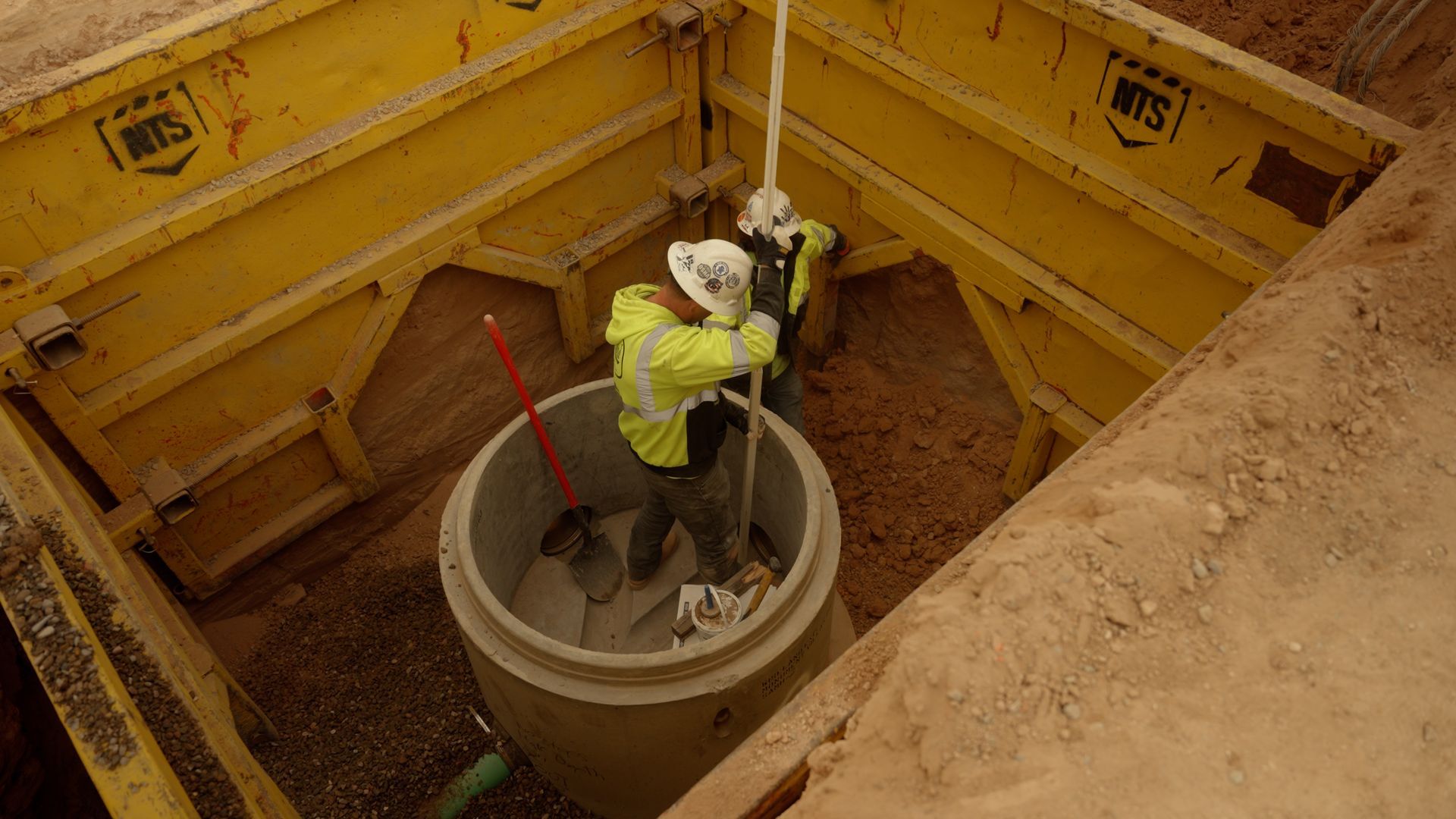 Worker in yellow safety gear standing in a concrete manhole within a dirt excavation.