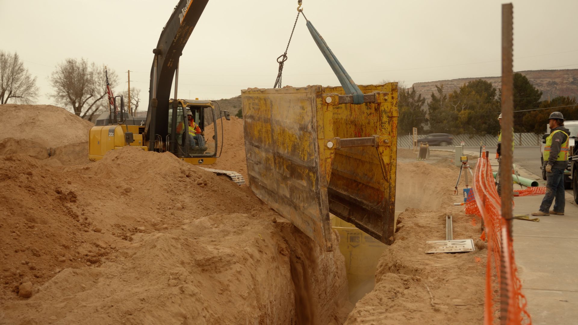Construction site with excavator lifting a yellow trench shield beside an orange safety fence