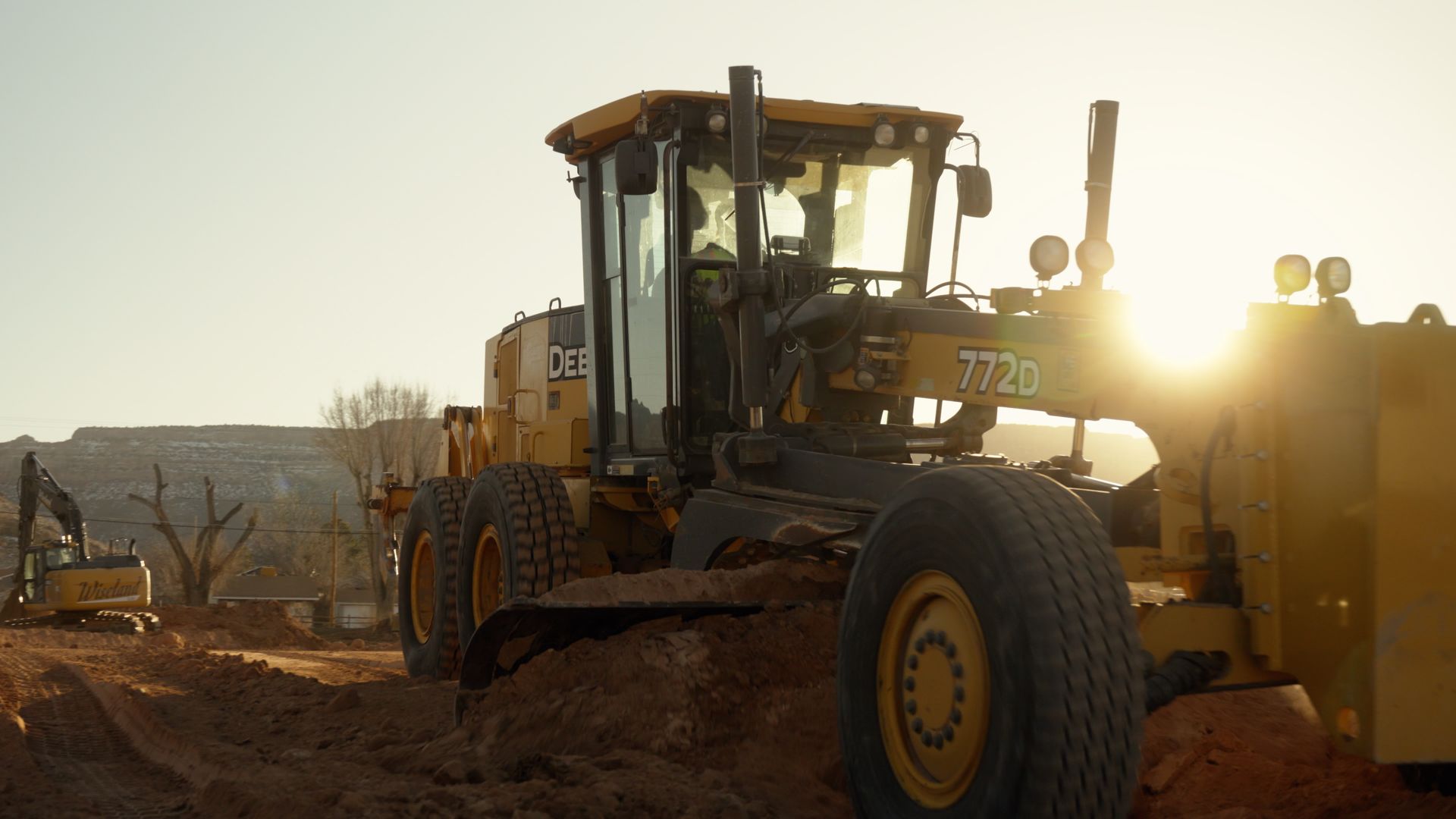 Yellow construction grader on dirt road at sunset, sun glare in the background