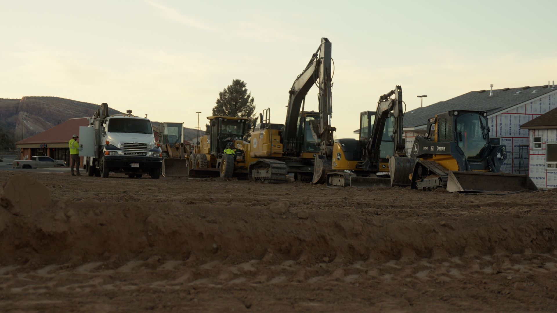 Construction equipment and trucks lined up on a dirt lot at dusk with houses in the background
