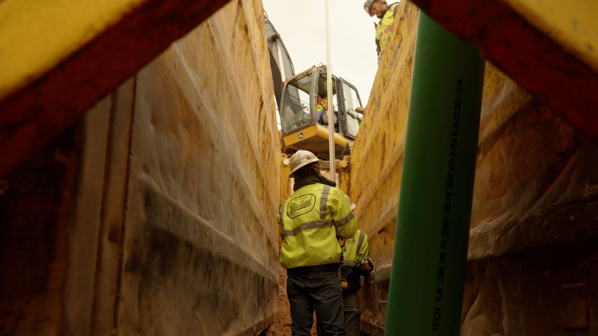 Worker in a hard hat and yellow safety vest standing in a narrow industrial trench between large metal walls.