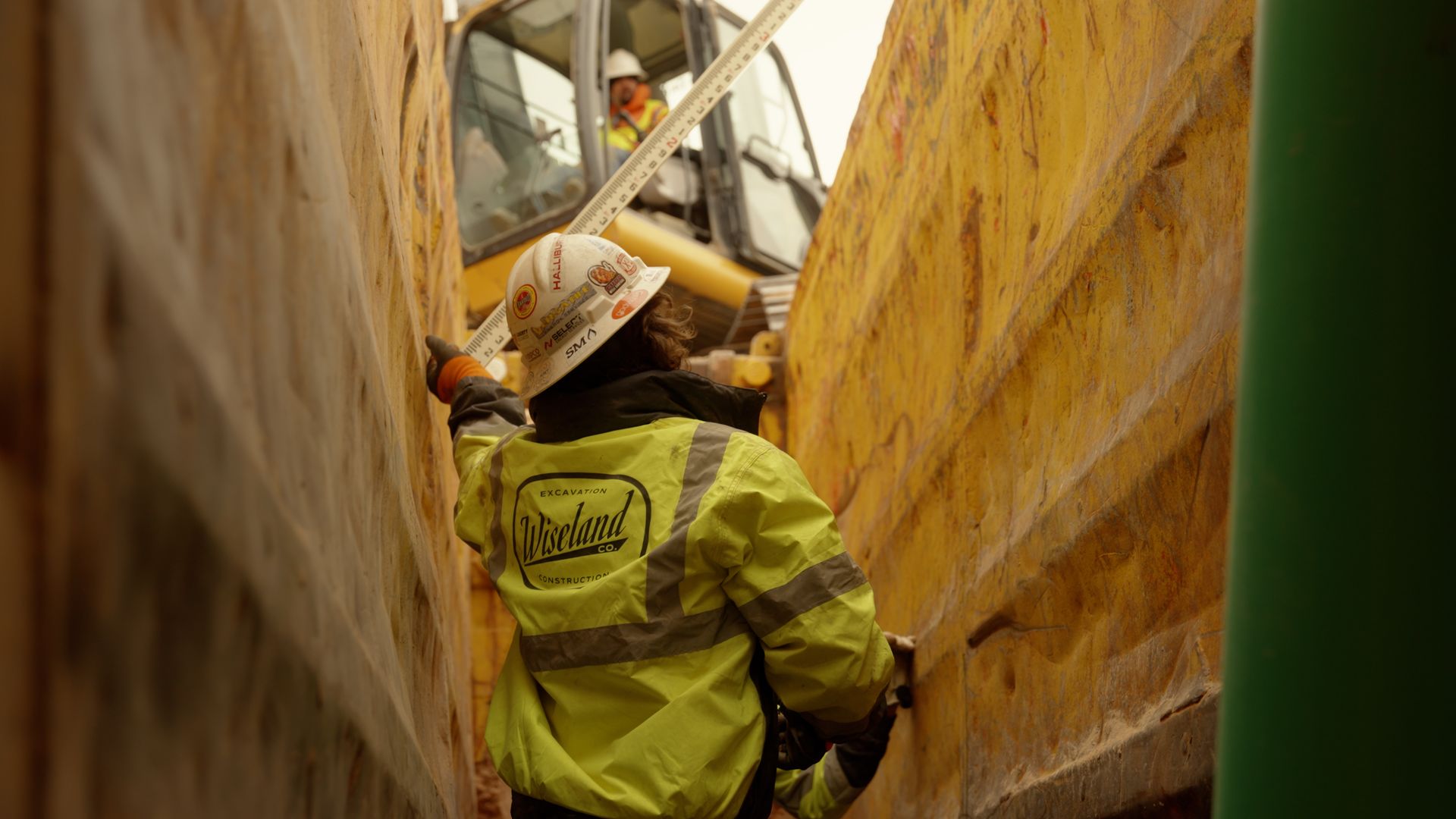 Worker in a yellow safety jacket and hard hat standing between tall wooden walls at a construction site.