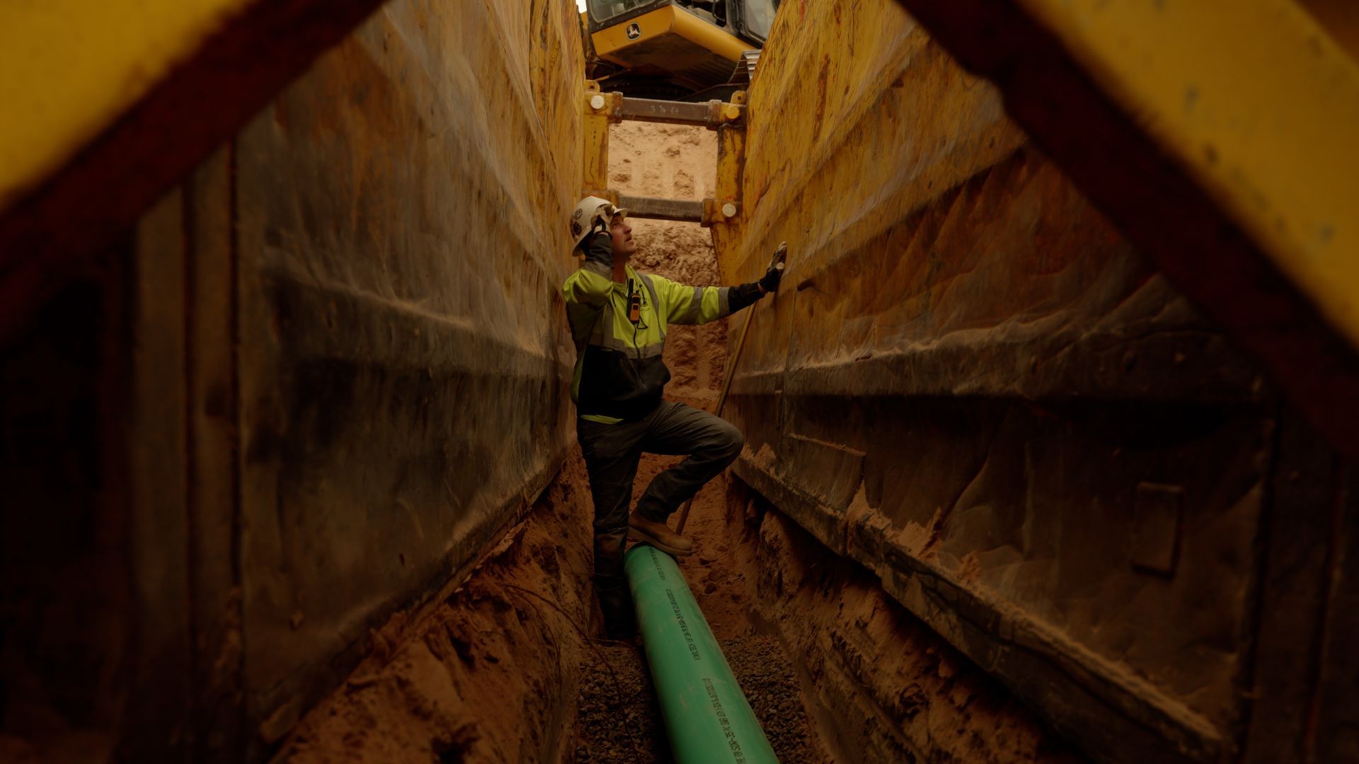 Worker in a narrow muddy trench, reaching over a green pipe between rust-colored walls.