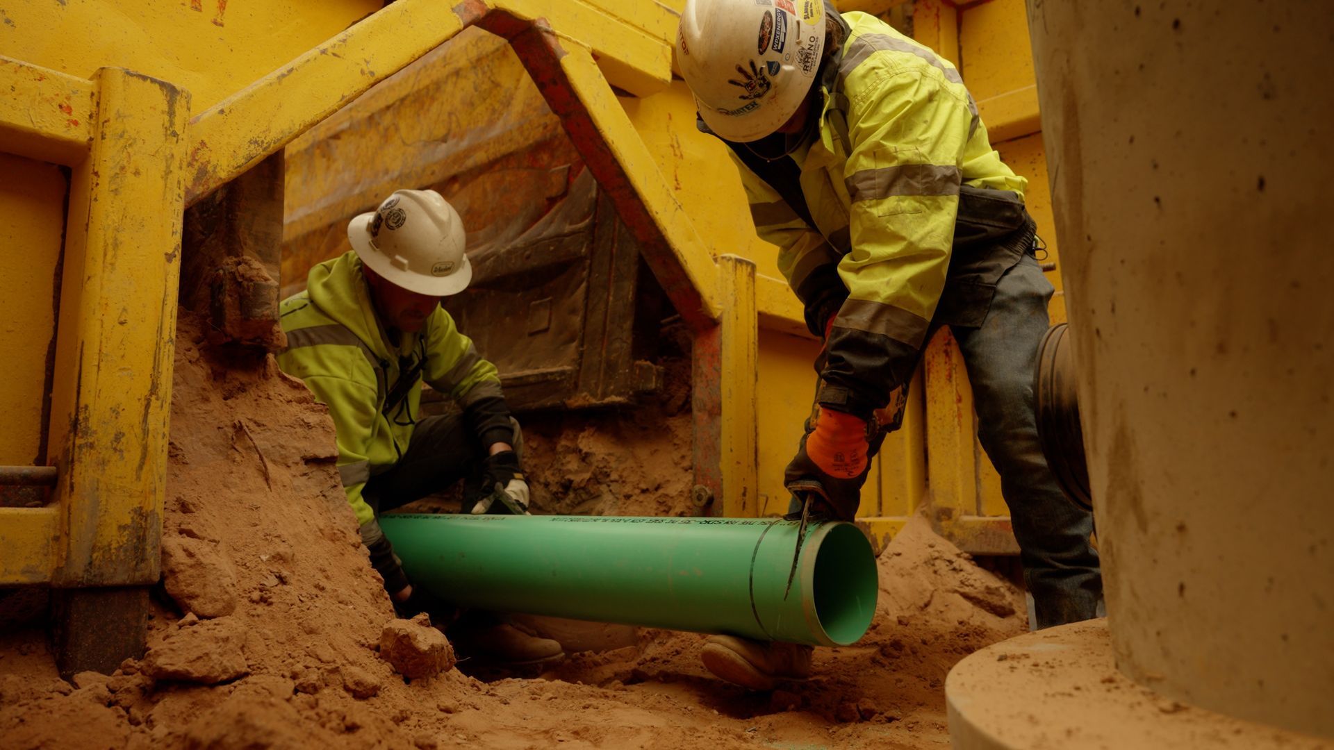 Construction workers installing a green pipe in a muddy trench under yellow shoring