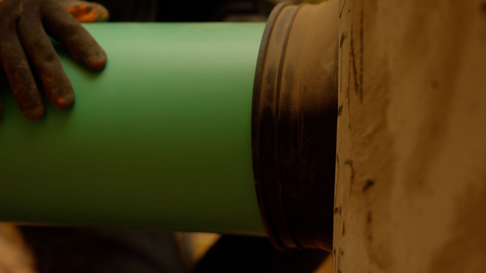 A hand holds a green pipe being fitted into a brown cylindrical pipe joint.