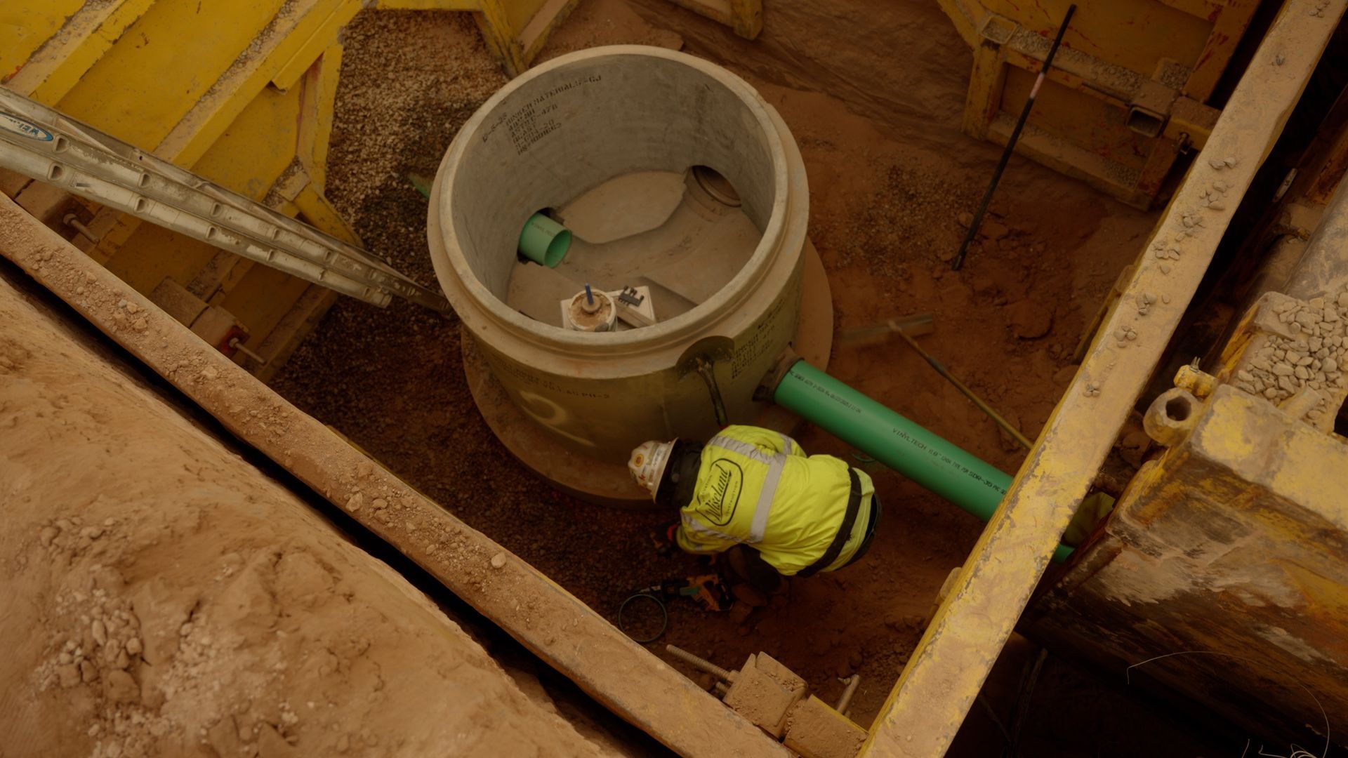 Construction trench with a concrete pipe chamber, green pipe, and yellow valve fitting amid wooden framing