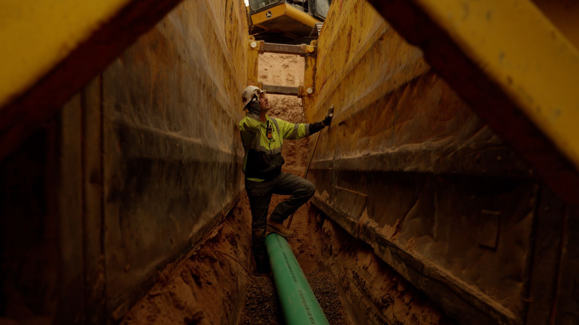 Worker standing in a narrow industrial trench beside a green pipe, with rusty metal walls overhead