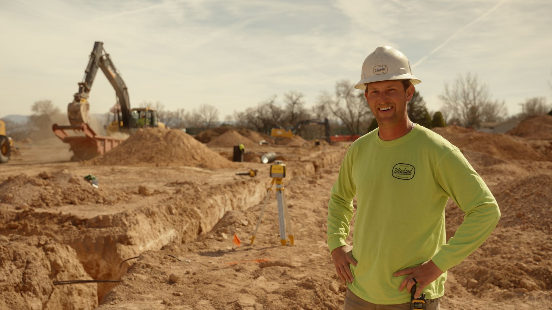 Construction worker in hard hat standing at a dirt excavation site with an excavator in the background