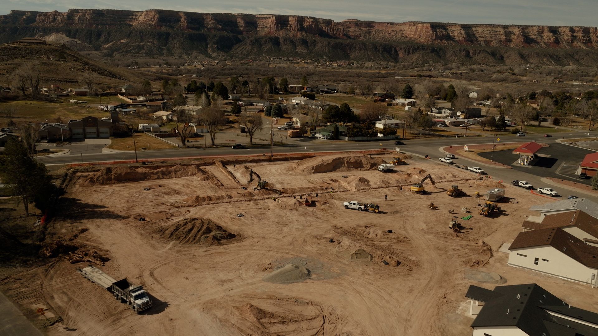 Construction site with excavated dirt, heavy equipment, and buildings in a desert town with mesas in the background