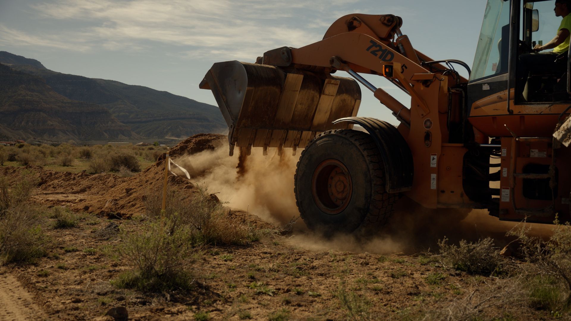 Orange excavator digging dusty soil in a dry, rocky field with hills in the background