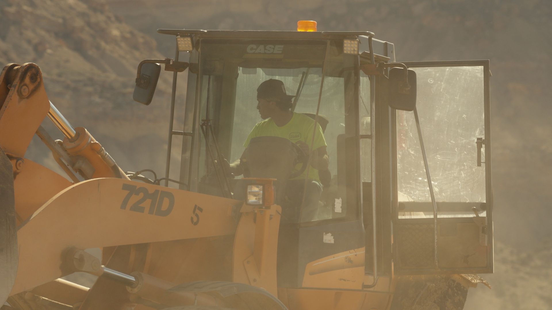 Yellow excavator in a dusty site with a worker visible inside the cab