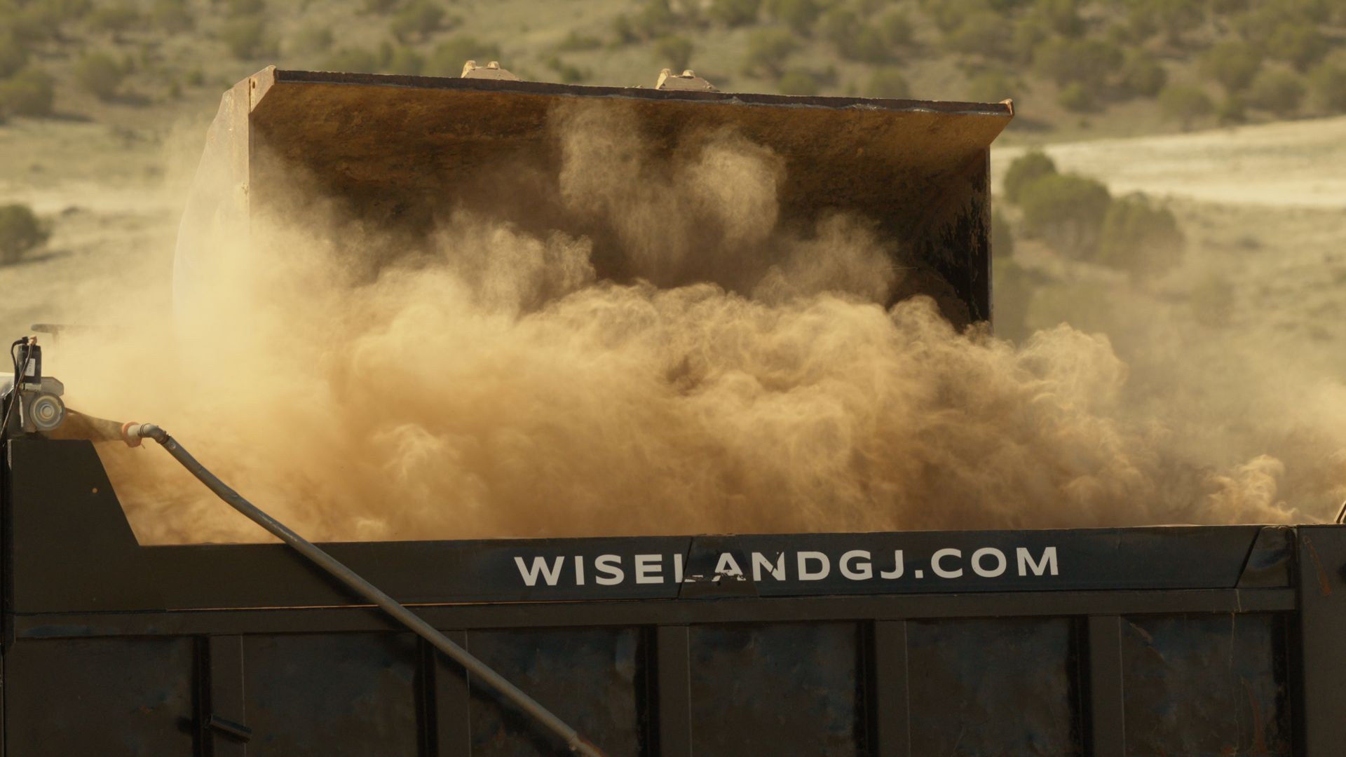 Dump truck bed raising a cloud of dust in a dry field, with wiselandgj.com on the side.