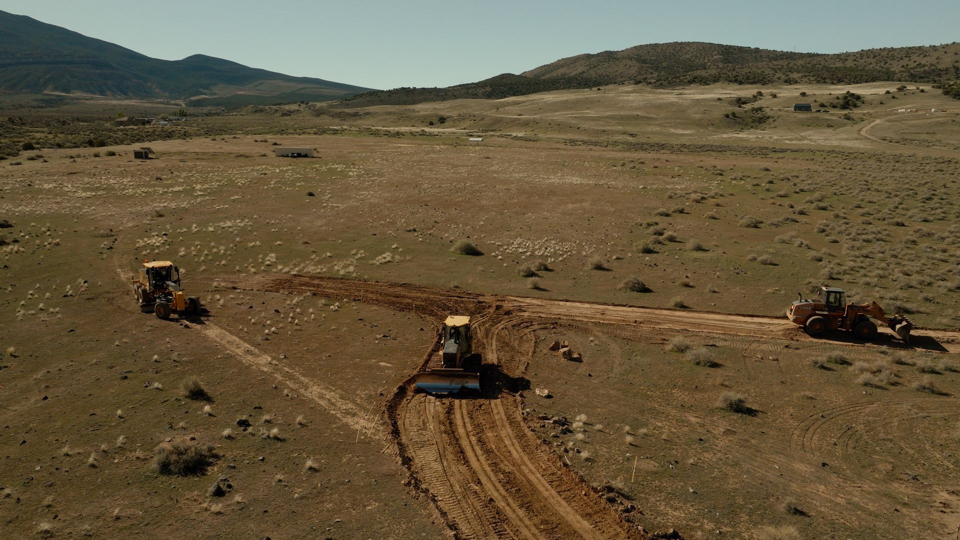 Three tractors work in a dry, rocky field with mountains in the background.