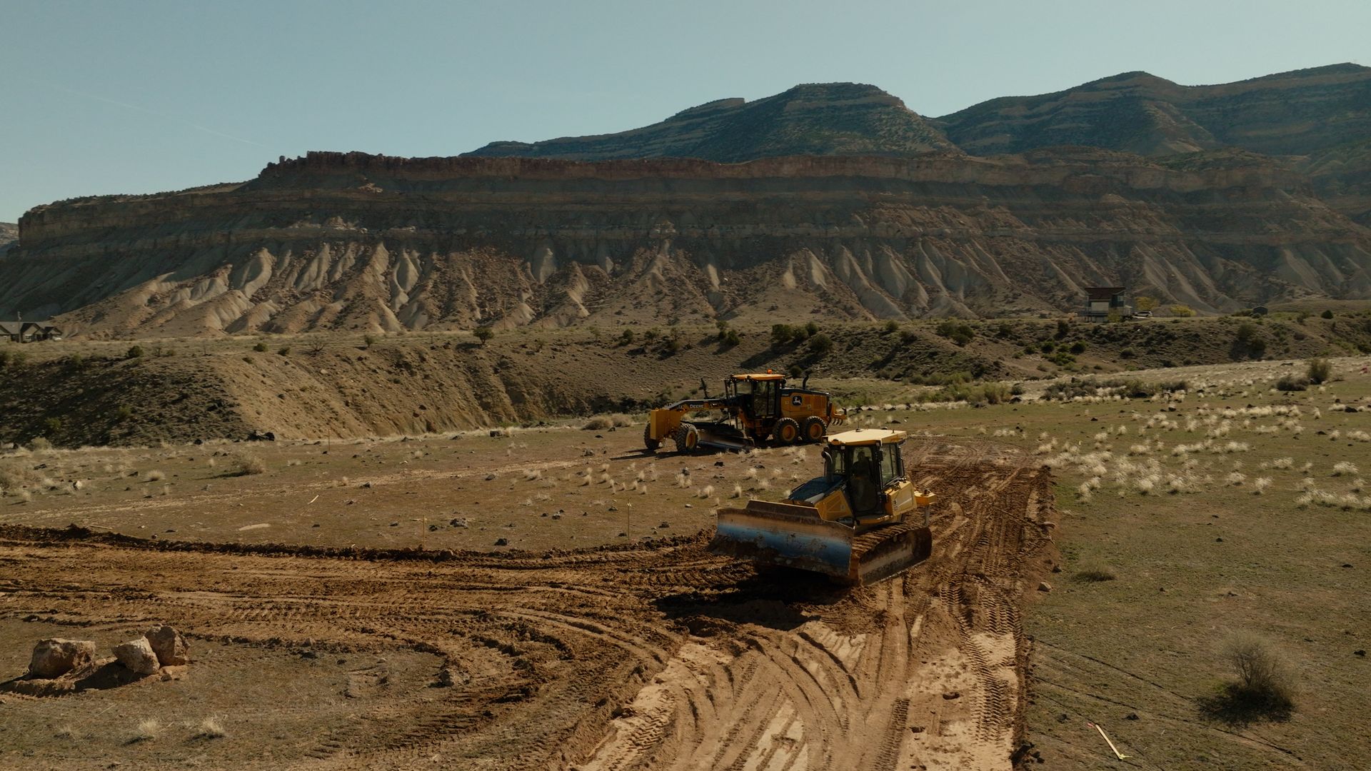 Bulldozer and construction vehicle working on a dirt road in a barren desert landscape with rocky hills.