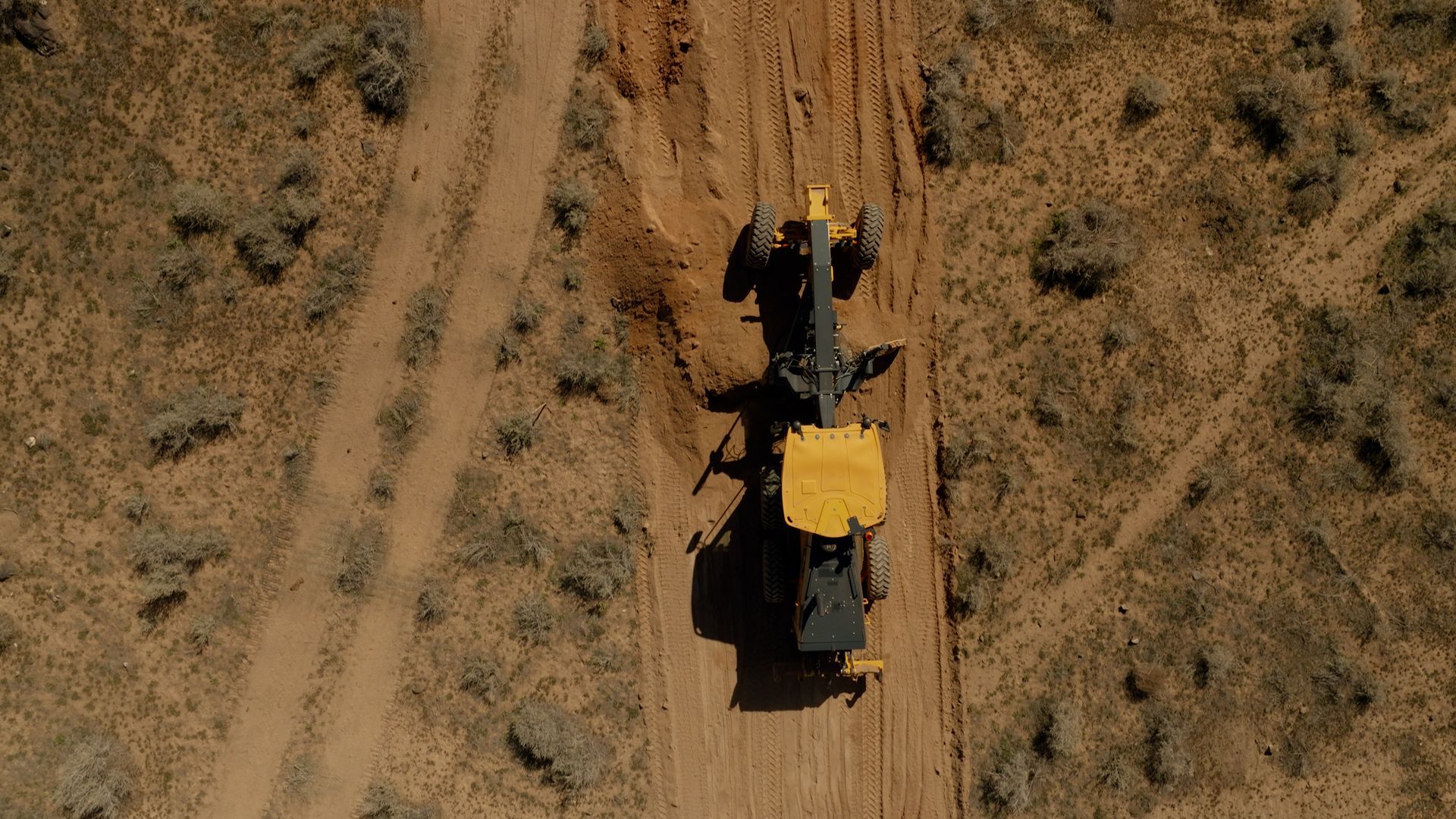 Overhead view of a yellow construction vehicle driving on a dirt road through dry scrubland