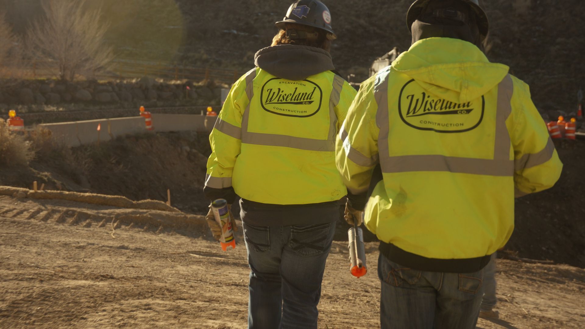 Two workers in neon yellow jackets stand near a construction site at dusk.