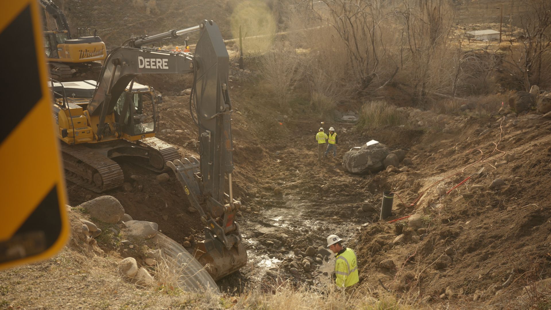 Excavator and workers in high-visibility vests at a muddy construction site beside a ditch