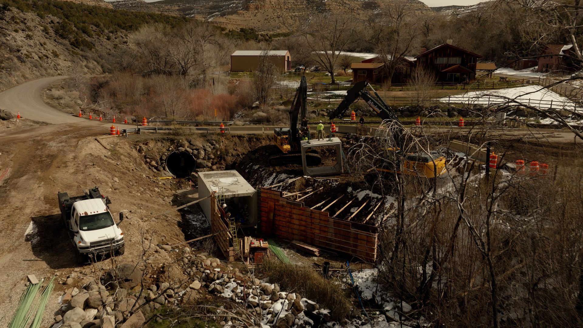 Damaged homes and debris in a wildfire-scarred neighborhood, with a white SUV on a dirt road